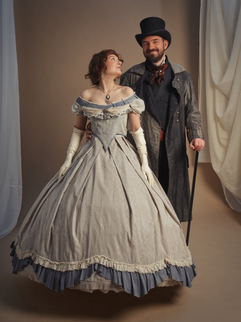 Miss Jack porte une robe Second Empire dans les tons de bleu. À côté d'elle, Romain porte un haut de forme, noire, un manteau foncé et une canne? 