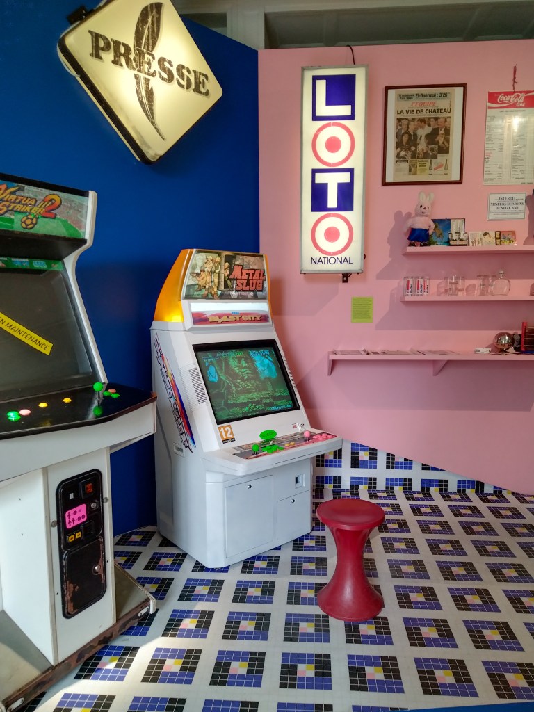 Deux bornes, dont l'une de Metal Slug. Un mur est bleu, l'autre est rose. Au sol, un motif en mosaïque. Sur les mieux, un néon "presse" et "Loto" et des babioles (peluche de lapin Duracel, une de journal, tarifs des consommations...).