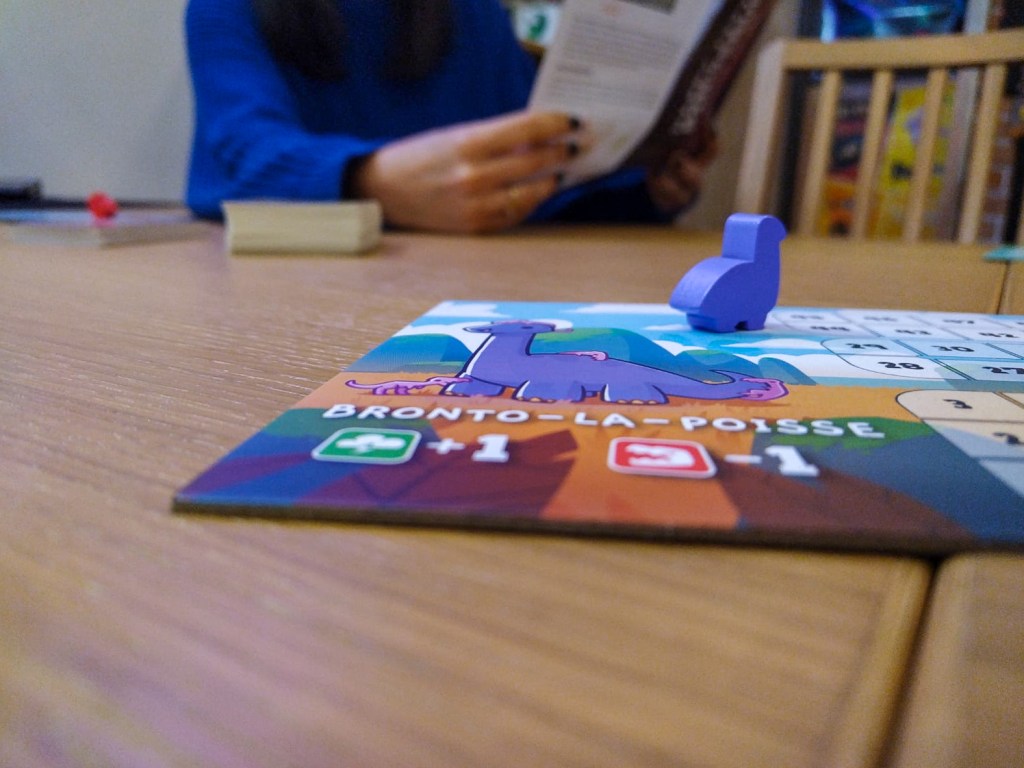 Partie d'un jeu de société où l'on incarne des dinosaures. Un petit diplodocus violet est sur une plaquette intitulée "Bronto la Poisse"